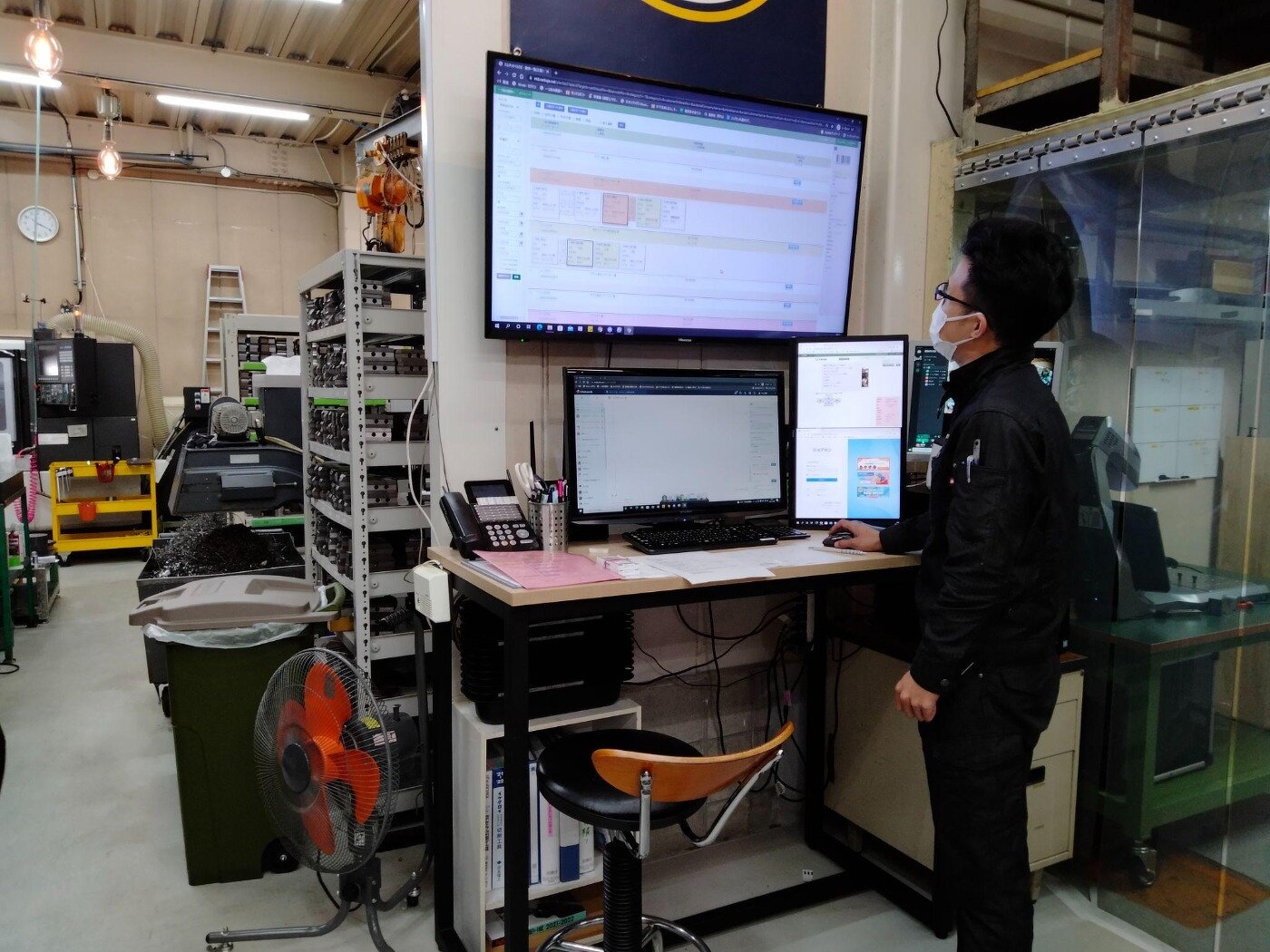 製造現場の壁面に設置された大型モニターで、エムネットくらうどの工程管理画面を確認する作業服姿の男性