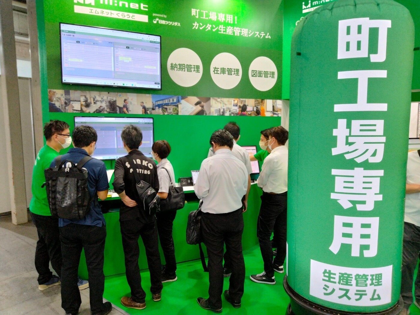 展示会ブースで生産管理システム「エムネットくらうど」の画面を囲み、説明を聞く来場者たち
