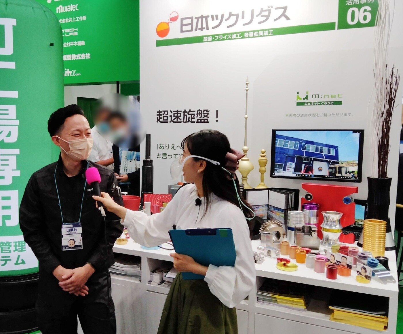 展示会ブースでインタビューを受ける日本ツクリダスの担当者と、カウンターに並ぶ金属加工製品の展示
