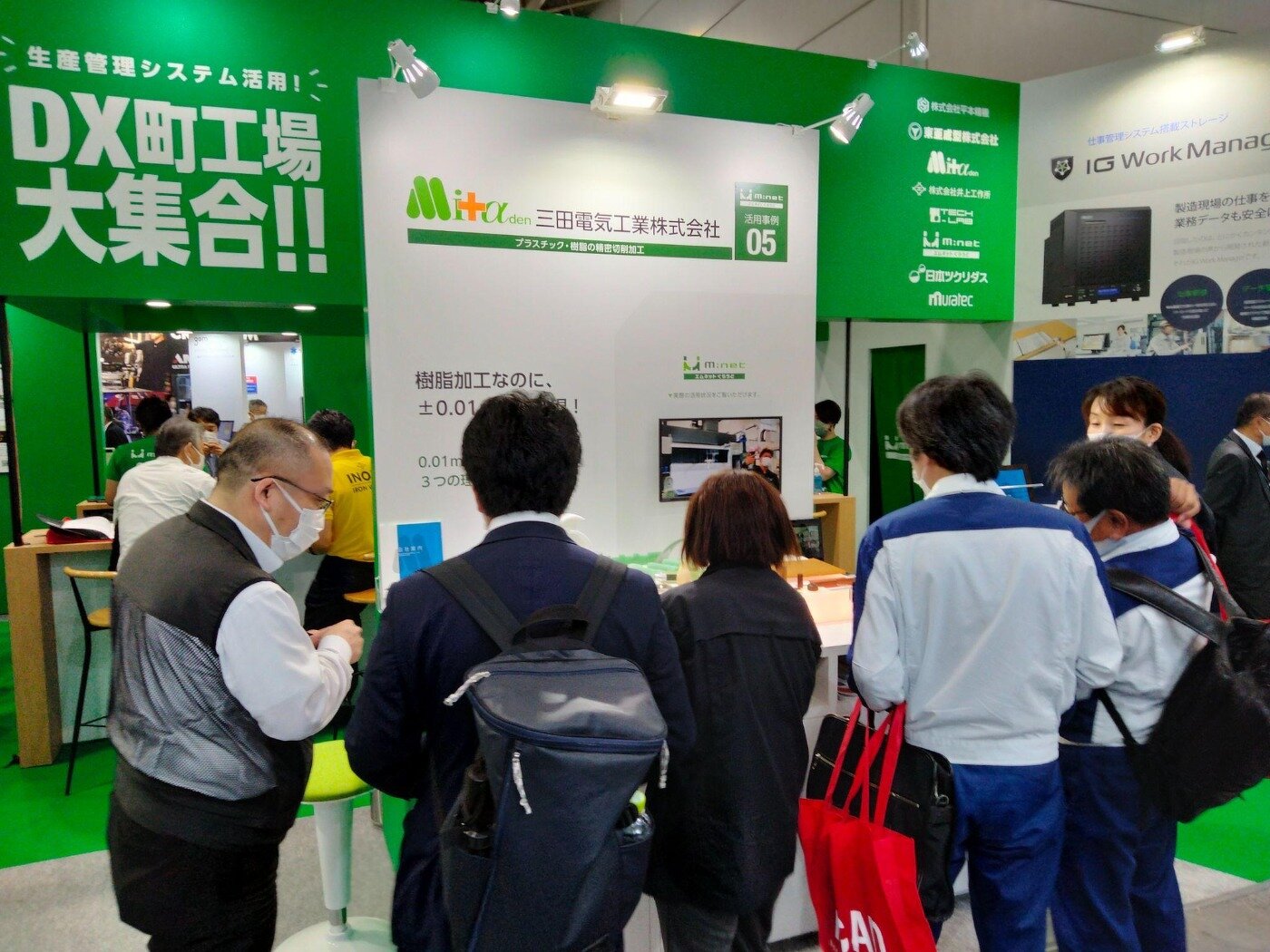 展示会ブースで展示パネルやモニター画面を見学する来場者と、説明を行う三田電気工業株式会社の担当者